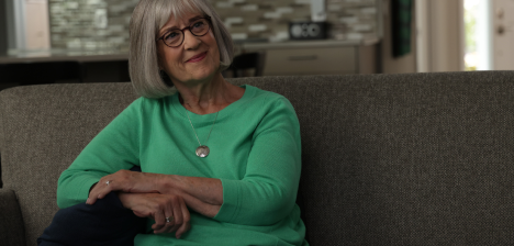 A patient story video of Mary, an adult female ABECMA® (idecabtagene vicleucel) patient, shown with glasses and short grey hair wearing a green long sleeve shirt while sitting on a couch with her arms crossed